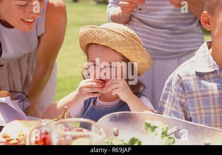 Parkszene, Porträt, 3 Generationen einer Familie geniessen einen Sommertag im Grünen bei einem Picknick, 6-jaehriges Maedchen mit Strohhut lutscht ein einer Erdbeere Stockfoto