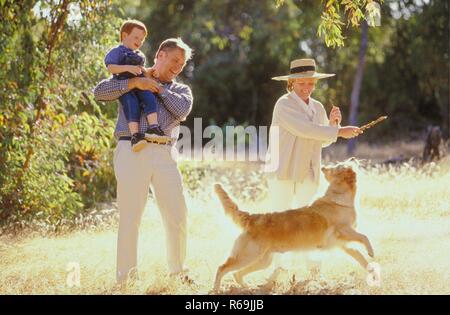 Porträt, Szene, Grossvater traegt-rothaarigen 3 Jahre alten Enkelsohn mit dem Arm, waehrend die Grossmutter mit ihrem Hund auf einer Wiese am Waldrand, mit einem Lager spielt Stockfoto
