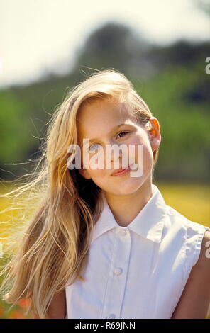 Porträt, laechelndes blondn Maedchen mit langen Haaren, 12 Jahre, mit weisser aermellosen Bluse sitzt in einer mit Klatschmohn und gelben Blueten uebersaeten Wiese Stockfoto