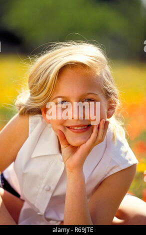 Porträt, laechelndes blondn Maedchen mit langen Haaren, 12 Jahre, mit weisser aermellosen Bluse sitzt in einer mit Klatschmohn und gelben Blueten uebersaeten Wiese Stockfoto