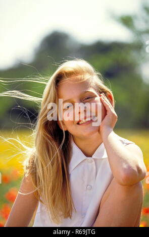 Porträt, laechelndes blondn Maedchen mit langen Haaren, 12 Jahre, mit weisser aermellosen Bluse sitzt in einer mit Klatschmohn und gelben Blueten uebersaeten Wiese Stockfoto