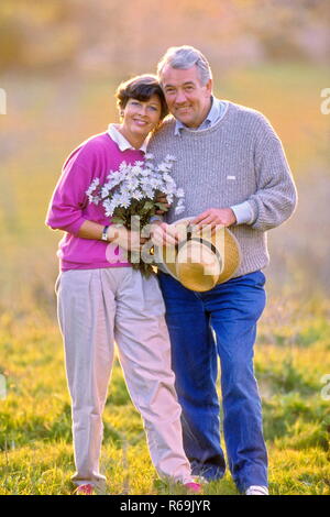 Seniorenpaar, Mann mit Jeans und Pullover und Frau mit weisser Schlauch und himbeerfarbener Strickjacke mit einem Strauß Margeriten und Strohhut in der Hand auf Wiese Stockfoto