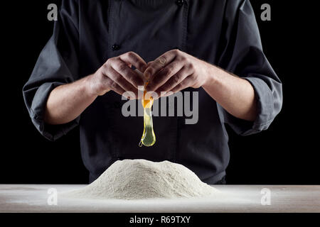 Fallende Eier in Mehl. Hände brechen, reißen ein rohes Ei über Mehl, Backpulver Konzept. Baker knetet den Teig für die Pasta. Der Prozess der Aufschlagen der Eier. Stockfoto