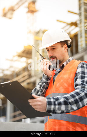 Entwicklungsingenieur tragen weiße Warnweste und Schutzhelm mit Walkie talkie und Zwischenablage Inspektion Baustelle. Entwicklung und Konstruktion Stockfoto