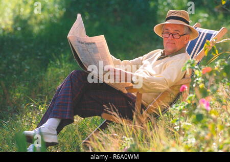 Portrait, Senior in Freizeitkleidung mit Brille und Strohhut sitzt im Garten im Liegestuhl und liest seine Zeitung Stockfoto