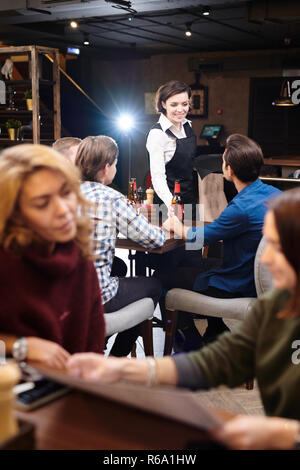 Positiven Inhalt schöne Dame Kellnerin in schwarzer Schürze steht am Tisch und die Flasche Bier zu den Männern beim freundlichen Gespräch mit Besuchern im Pub Stockfoto
