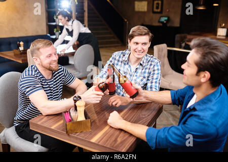 Gruppe von Jolly begeistert Junge männliche Freunde in Freizeitkleidung sitzt am Tisch in der Bar und Toasten mit Bierflaschen mit Spaß am Freitag Stockfoto