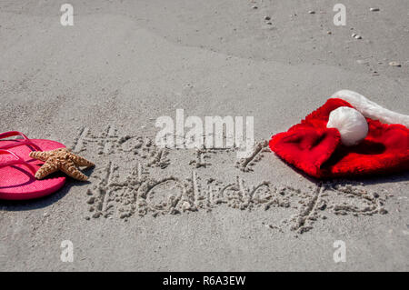 Tropische Weihnachten am Strand, mit Happy Holidays in den Sand geschrieben, Flip Flops, ein Seestern und Santa hat. Stockfoto