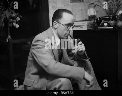 1950, historische, ein Mann im Anzug sitzt das Rauchen einer Pfeife in Innenräumen zu Hause, England. Pfeife rauchenden war zu dieser Zeit sehr beliebt. Auch auf dem Bild eine Bakelit GPO Telefon der ära gesehen. Stockfoto