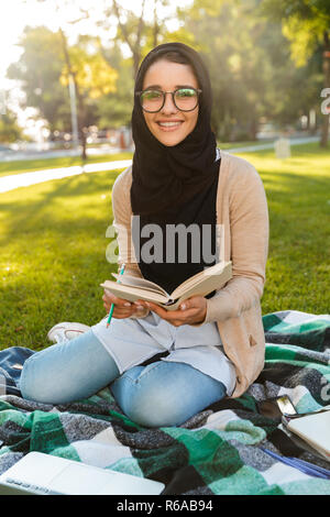 Foto von attraktiven muslimische Frau mit Kopftuch sitzt auf Decke im Green Park und lesen Buch Stockfoto