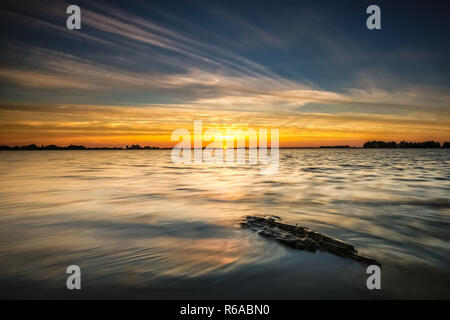 Sonnenuntergang an der Kante entlang der Seen in Flevoland. Floating Stück Treibholz an der Küste mit Blick über die flache holländische Polderlandschaft gewaschen. Stockfoto