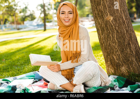 Foto der schönen arabischen Frau mit Kopftuch sitzen auf Decke im Green Park und lesen Buch Stockfoto