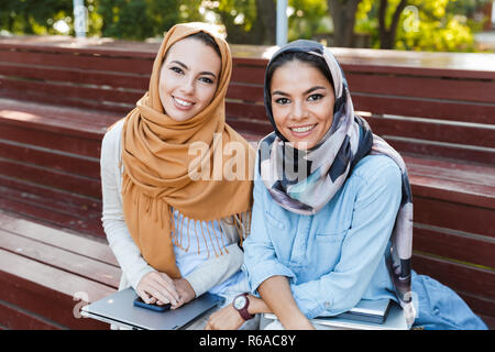 Foto von zwei muslimischen Frauen das Tragen von headscarfs Lächeln auf die Kamera, während auf der Bank im Park sitzen Stockfoto