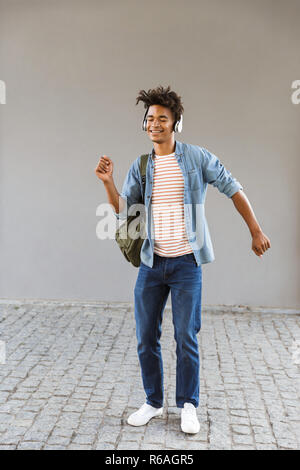 Freudige junge afrikanische Mann mit Rucksack im Freien, Musik hören mit Kopfhörern, Tanzen Stockfoto