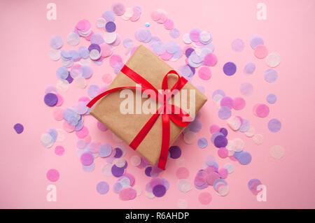 Geschenkbox in braun gefärbte Handwerk Papier gewickelt und mit roter Schleife auf rosa Hintergrund mit colofrul Konfetti gebunden. Stockfoto