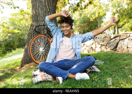 Fröhlicher junger afrikanischer Jugendlicher mit Fahrrad im Freien, im Gras sitzen, Musik hören mit Kopfhörern, Spaß, Tanzen Stockfoto