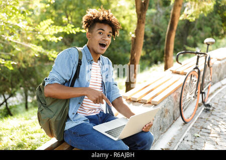 Bild von schockiert stattlichen emotionale Junge afrikanische Kerl in Park im Freien mit Laptop Computer. Stockfoto