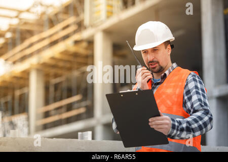 Entwicklungsingenieur tragen weiße Warnweste und Schutzhelm mit Walkie talkie und Zwischenablage Inspektion Baustelle. Entwicklung und Konstruktion Stockfoto