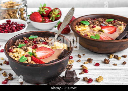 Schokolade Banane protein Smoothie Schüsseln mit Müsli, Erdbeere, Granatapfel und Schokolade zum Frühstück auf weiße Holztisch serviert. nach oben schließen. Stockfoto