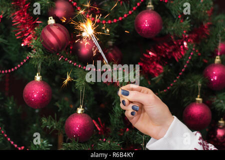 Mädchen Hand, die brennende Wunderkerze blast und Weihnachtsbaum mit roten Dekoration in den Hintergrund. Weihnachten und Neujahr Konzept. Nahaufnahme Stockfoto