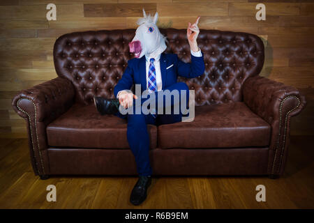 Lustige Einhorn im eleganten Anzug Standortwahl auf dem Sofa wie ein Chef und zeigt Geste mit erhobenem Zeigefinger. Ungewöhnliche Mann zu Hause. Freaky Kerl in der Maske. Stockfoto