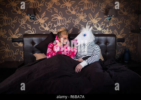 Junge Frau im Pyjama sitzt auf dem Bett und spricht mit lustigen Freund in komischen Einhorn Maske. Ungewöhnliche Paar verbringt die Zeit zusammen in einem stilvollen Schlafzimmer. Stockfoto
