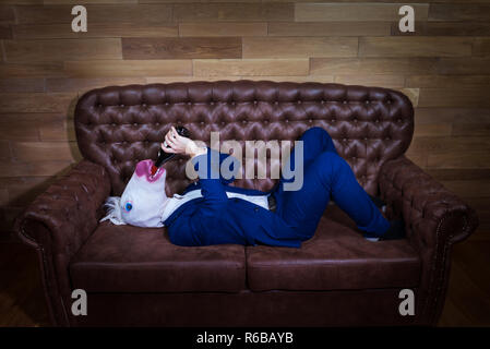 Lustige Einhorn im eleganten Anzug liegt auf einem Sofa. Ungewöhnliche mann Getränke Wein aus der Flasche zu Hause. Freaky Chef in komischen Maske auf dem Hintergrund der hölzernen Wand. Stockfoto