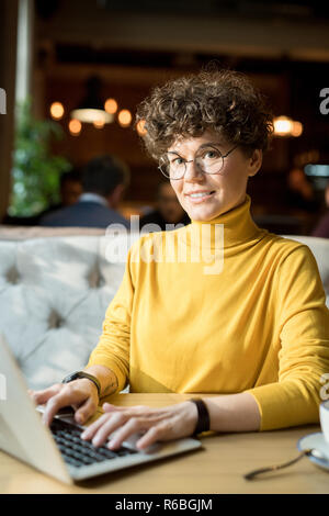 Fröhliche schönen lockigen Haaren hippie Frau mit Brille gelb Rollkragen sitzen am Tisch und Arbeiten mit Laptop, während bei Kamera suchen Stockfoto