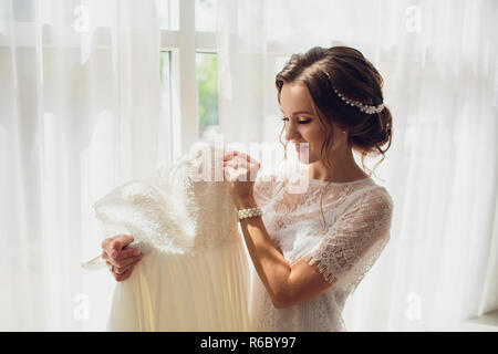 Hübsche brünette Braut in Seide Morgenmantel und Lacy Schleier Holding Hochzeit Kleid auf einem Kleiderbügel in der Nähe der Fenster. Stockfoto