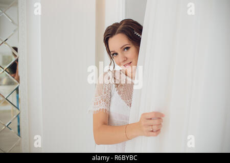 Hübsche brünette Braut in Seide Morgenmantel und Lacy Schleier Holding Hochzeit Kleid auf einem Kleiderbügel in der Nähe der Fenster. Stockfoto