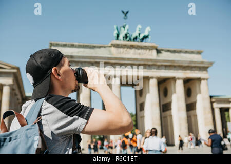 Eine Person, die Getränke aus einer Tasse Kaffee oder einem anderen Getränk bei einem Spaziergang oder beim Sightseeing in Berlin in Deutschland. Die Zukunft ist das Brandenburger Tor und verschwommenes unkenntlich Menschen. Stockfoto