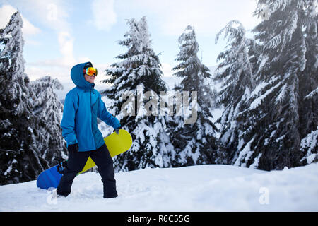 Snowboarder, Klettern, Mountain Top seinem Snowboard tragen durch Wald für backcountry Freeride und Tragekomfort Reflektierende Schutzbrille, bunte Mode Outfit und Sturmhaube bei Ski Resort. Wintersport. Stockfoto