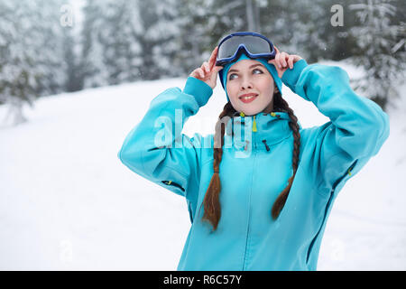 Lächelnd nordic Frau mit Zöpfen setzt an schützenden Skibrillen. Snowboarder Mädchen berühren Maske bei Ski Resort auf Schneefall in der Nähe von Forest. Blue eyed elegante Sportlerin in bunten modische Outfit. Stockfoto