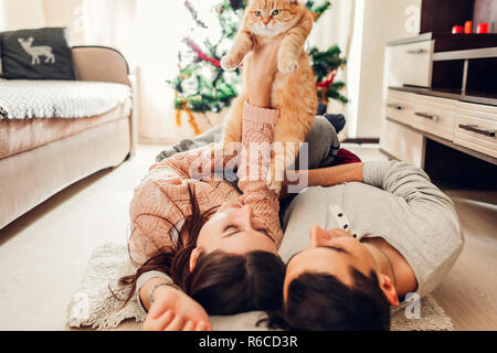 Verliebtes Paar liegen, Weihnachtsbaum und das Spielen mit der Katze zu Hause. Mann und Frau Heben von Pet in den Händen. Glückliche Familie Stockfoto