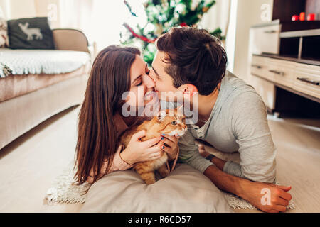 Verliebtes Paar liegen, Weihnachtsbaum und das Spielen mit der Katze zu Hause. Glücklich der Mann und die Frau küssen