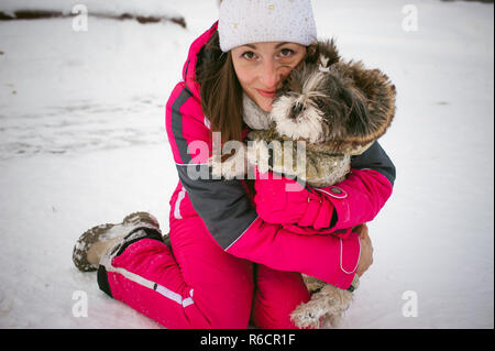 Spaziergang im Winter im Freien mit Hund Rasse Shih Tzu. Eine Frau in leuchtend roten warme Skibekleidung Wandern im Schnee mit Ihrem Haustier, kleine Shih Tzu in Overalls gekleidet. Pflege für Tiere liebt das Spiel mit dem Hund Stockfoto