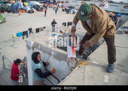 Tijuana, Mexiko. 04 Dez, 2018. Ein Wanderarbeitnehmer hilft seinen Neffen eine Mauer klettern. Im Hintergrund sind Zelte für die Migranten in der mexikanischen Stadt Tijuana warten. Derzeit mehr als 6000 Migrantinnen aus Mittelamerika warten in der Grenzstadt mexikanischen Tijuana Asyl in den USA zu bewerben. Da nur eine begrenzte Anzahl von Fällen im San Ysidro Grenzübergang verarbeitet werden, es gibt Monate Wartezeit. Credit: Bruno Gallardo/dpa/Alamy leben Nachrichten Stockfoto