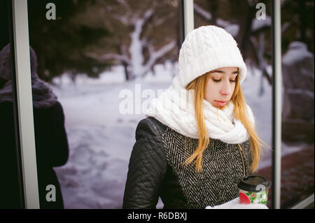 Frau in weißen Schal und Hut trinkt Kaffee. cute blonde junge Frau, die in der Nähe von Spiegelbild Vitrinen Cafe, Kaffee trinken zu gehen, mit den pappbecher in der Hand Stockfoto