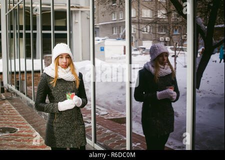 Frau in weißen Schal und Hut trinkt Kaffee. cute blonde junge Frau, die in der Nähe von Spiegelbild Vitrinen Cafe, Kaffee trinken zu gehen, mit den pappbecher in der Hand Stockfoto