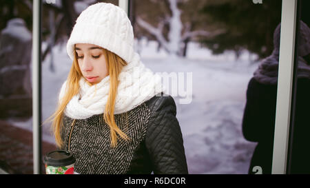 Frau in weißen Schal und Hut trinkt Kaffee. cute blonde junge Frau, die in der Nähe von Spiegelbild Vitrinen Cafe, Kaffee trinken zu gehen, mit den pappbecher in der Hand Stockfoto