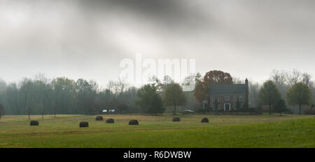 Heuballen in Feld mit Haus im Hintergrund am nebligen Morgen Stockfoto