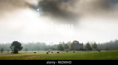 Heuballen in Feld mit Haus im Hintergrund am nebligen Morgen Stockfoto