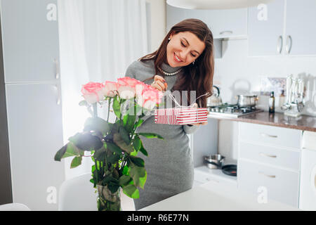 Junge Frau gefunden Geschenkbox und Blumenstrauß aus Rosen auf Küche. Glücklich lächelnde Mädchen anwesend Stockfoto