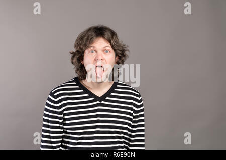 Portrait von lustigen shaggy verrückt gutaussehenden Mann im gestreiften Pullover bei Kamera schaut mit großen offenen Mund und Zunge, quält. Die weit geöffneten Augen indoor Studio shot, auf grauem Hintergrund. Stockfoto