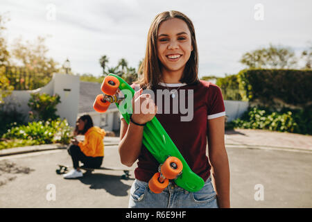 Lächelnd Teenager auf der Straße stand ihr skateboard Holding mit ihrer Freundin Eislaufen im Hintergrund. Junge Mädchen üben Eislaufen in der St Stockfoto