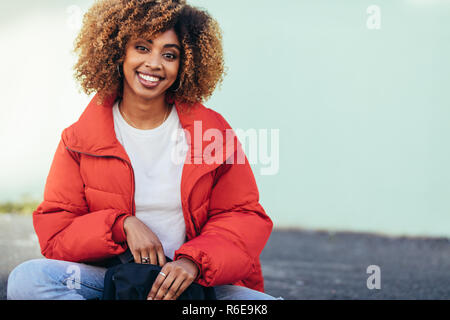 Fröhliche afro-amerikanische Frau sitzt draußen auf der Straße. Lächelnd touristische Frau in Jacke sitzen auf der Straße eine Tasche. Stockfoto