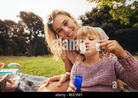 Süße kleine Mädchen bläst Seifenblasen beim Sitzen mit ihrer Mutter im Freien. Mutter und Tochter Spaß an Picknick im Park. Stockfoto
