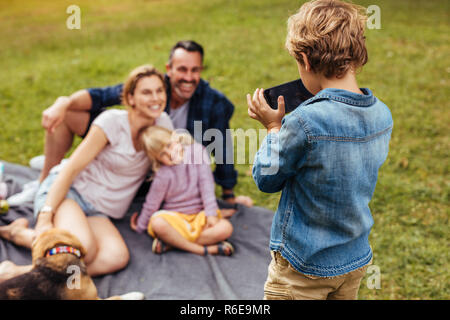 Little Boy unter Foto von seinen fröhlichen Familie sitzt auf der Decke im Park. Sohn fotografieren Familie während Picknick im Park. Stockfoto