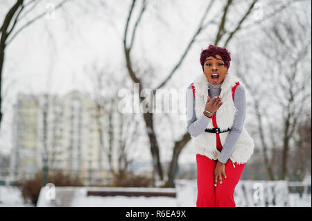 Afrikanische amerikanische Frau in roten Hosen und weißen Pelz Jacke stellte auf der Winter Tag gegen verschneite Hintergrund, zeigt eine überrascht Emotion. Stockfoto