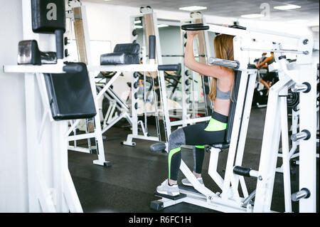 Mädchen in einem hellen Weiß Fitnessstudio. Eine blonde Frau eine Stärke Übung für eine Gruppe von Muskeln auf die Hände eines Simulators. Gesunde Lebensweise, guter Laune Stockfoto